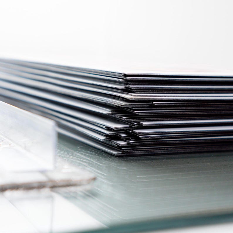 May include: A stack of rectangular, black and white cards or papers, possibly photographs or prints, arranged in a neat pile. The edges of the cards are visible, creating a striped effect. The surface is reflective, with a blurred object in the foreground.