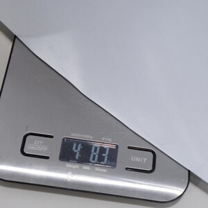 May include: A digital kitchen scale with a silver finish displays a weight of 48.3 ounces. The scale has an on/off button and a unit button. The maximum weight capacity is 3000 grams. The scale is partially covered by a white sheet of paper.