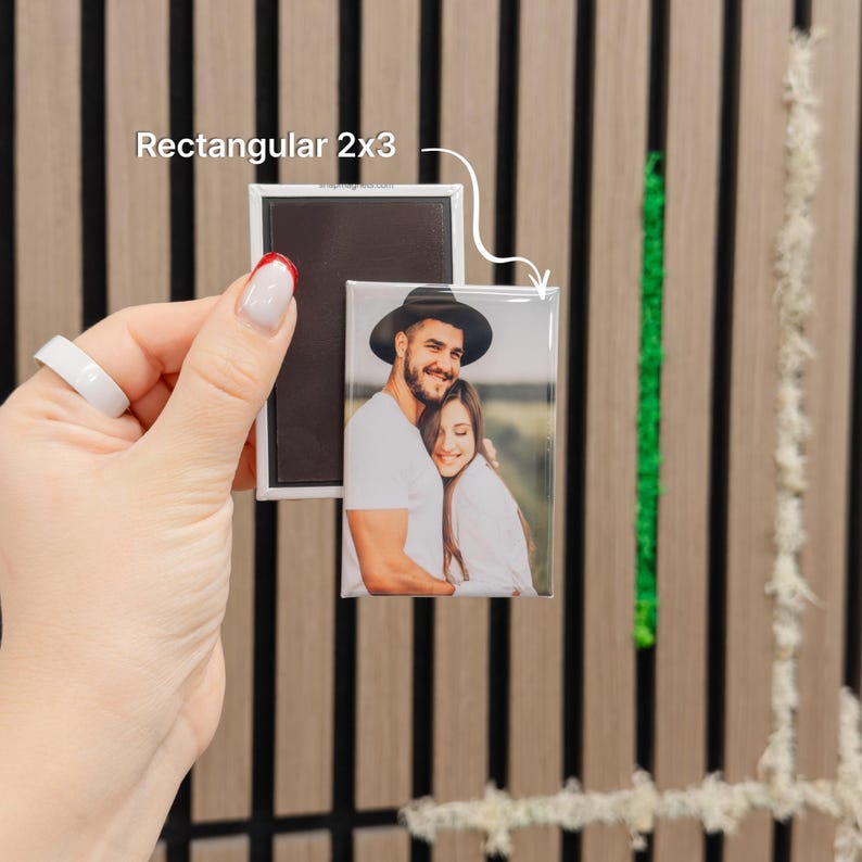 A hand holding a rectangular magnet with a photo of a couple on it, against a wooden background with vertical lines.