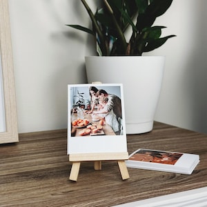 May include: A small wooden easel displaying a rectangular photograph of a family preparing food with oranges on a table. The photograph features a white border and is positioned upright on the stand, with a stack of identical photographs nearby.