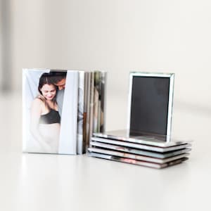 May include: A stack of square photo magnets with a black magnetic surface and a silver frame. A stack of square photo prints is next to the magnets. The photos show a pregnant person and their partner.