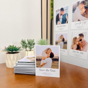 May include: A stack of rectangular photo cards featuring a couple embracing in a field. Each card displays the text Save the Date, Sam & Chloe, September 28, 2027, Wisconsin. The cards are positioned near two small green succulents in white pots.