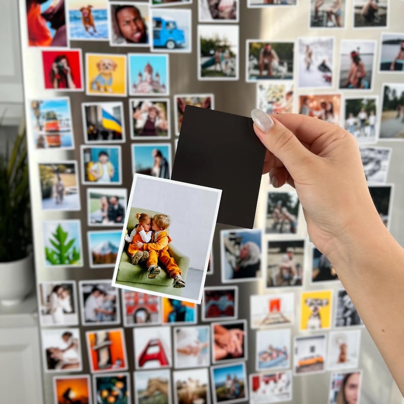 A hand is holding a small black magnet with a photograph of two people on it, in front of a refrigerator covered in various small magnets and photographs.