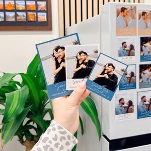 A hand holding three small blue and white wedding invitation cards with a couple posing together.