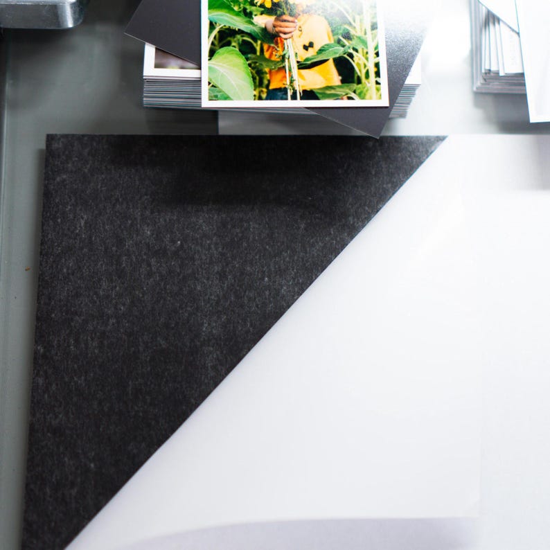 May include: A close-up shot shows a black, textured material in the upper left corner, meeting a white surface at a diagonal. A stack of small, dark photos and a larger photo with a person holding sunflowers are visible.