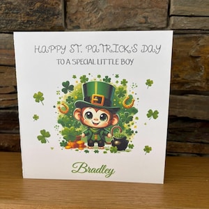 Può includere: Biglietto di auguri per San Patrizio con una scimmia dei cartoni animati con cappello a cilindro e abito verde, circondata da trifogli e un vaso d'oro. Il biglietto recita "Happy St. Patrick's Day To a Special Little Boy" con il nome "Bradley".