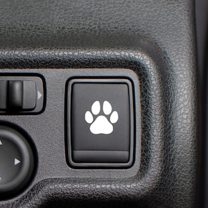 May include: A black car interior with a white paw print button. The button is located on the center console and is surrounded by other buttons.