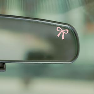 May include: A light pink bow decal applied to the surface of a vehicle rearview mirror. The bow is simple in design with two loops and two downward-pointing tails, positioned in the upper right corner of the reflective glass surface.