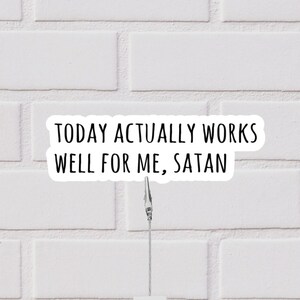 May include: A white sticker with black text that reads "TODAY ACTUALLY WORKS WELL FOR ME, SATAN." The sticker is held by a metal clip against a white brick wall background.