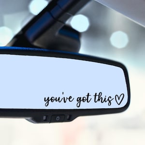 May include: A car rearview mirror with the phrase "you've got this" in black script, followed by a heart symbol. The mirror is set against a light blue background, with the car's interior visible.