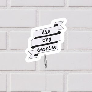 May include: A white sticker with a black banner design. The banner reads "die cry despise" in a bold, sans-serif font. The sticker is held by a silver metal clip against a white brick wall.