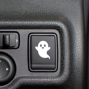 Puede incluir: Botón negro del interior de un coche con un icono de fantasma blanco. El fantasma tiene una cara sonriente y una cola curvada. El botón es rectangular y está situado en un panel negro texturizado.
