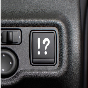 May include: Close-up of a black car interior panel featuring a button with a white exclamation point and question mark symbol. Other buttons with arrow symbols are also visible.