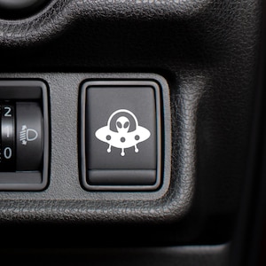 May include: A black car dashboard with a round knob labelled "0" and "2" and a square button with a white alien spaceship icon.