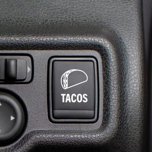 May include: Close-up of a black button with a white taco icon and the word "TACOS" in white text. The button is part of a car's interior control panel, surrounded by other buttons and a textured black surface.
