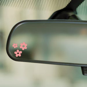 May include: A car's rearview mirror with three pink flower decals. The flowers are in a cluster, with two smaller flowers above a larger one. The mirror is black and has a reflective surface.
