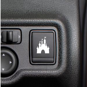 May include: A rectangular black plastic car dashboard button featuring a white silhouette graphic of a castle with multiple towers and flags. The button is set within a textured black plastic console panel alongside other vehicle control switches.