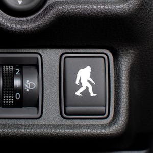 May include: Close-up of a black car dashboard with a light control dial and a button featuring a white Bigfoot silhouette. The dashboard has a textured surface.