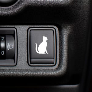 Op de afbeelding: Detail van een auto dashboard met een zwart paneel met een witte kat silhouet knop. Daarnaast een koplampbedieningsknop met instellingen 0, 1 en 2. Het dashboard heeft een getextureerd oppervlak.