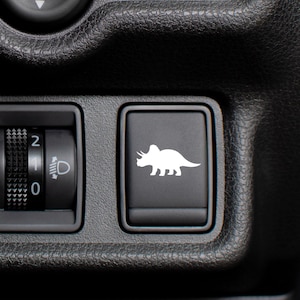 May include: Close-up of a black car interior panel featuring a white dinosaur graphic on a button. A headlight control dial is also visible. The panel has a textured surface.