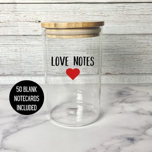 May include: A clear glass jar with a wooden lid, labeled "LOVE NOTES" with a red heart. A black circle on the left reads "50 BLANK NOTECARDS INCLUDED". The jar is on a white marble surface.