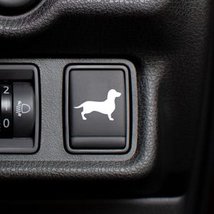Puede incluir: Primer plano de un salpicadero de coche negro con dos paneles de control. El panel izquierdo tiene un dial con los números 0, 1 y 2, y un símbolo de faro. El panel derecho tiene un icono de perro salchicha blanco.