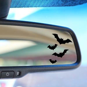 May include: Black bat decals are displayed on a car's rearview mirror. The bats are in various sizes and positions, creating a spooky Halloween theme. The mirror is set against a blue sky background.