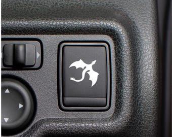 May include: A rectangular black plastic automotive switch featuring a white graphic of a dragon with wings spread. The switch is set into a textured black dashboard panel alongside other circular and rectangular control buttons for vehicle operation.