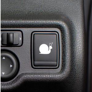 May include: A rectangular black plastic car dashboard button featuring a white graphic icon of a snail. The button is set within a textured black plastic console panel alongside other control switches with directional arrow symbols.