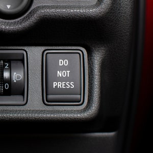 May include: A black car dashboard with a round knob and a square button. The knob has numbers 0, 1, and 2 on it. The button says "DO NOT PRESS".
