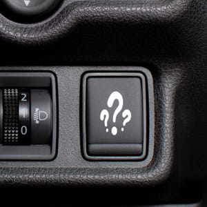 May include: A black rectangular vehicle dashboard button featuring a white graphic of three question marks arranged in a cluster. To the left, a black cylindrical dial with white numbers 0, 1, and 2 and a headlight icon is visible.