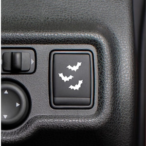 May include: A black car interior detail featuring a button with three white bat silhouettes. The button is part of a control panel with other buttons and a textured surface.