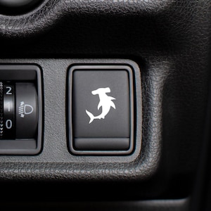 May include: A black rectangular vehicle dashboard button featuring a white silhouette of a hammerhead shark. To the left, a circular dial with numbers 0, 1, and 2 and a headlight icon is visible on the textured black plastic console surface.