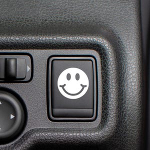 May include: Close-up of a black button with a white smiley face icon. The button is part of a larger black control panel, with other buttons and controls visible. The image has a focus on the smiley face.