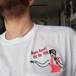 May include: White t-shirt with a pocket featuring an embroidered design. The design includes the text "Angela Bassett did the thing" and an illustration of a woman holding a microphone, dressed in pink.
