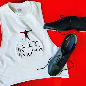 Puede incluir: Una camiseta sin mangas blanca con un diseño gráfico rojo y negro que presenta a un bailarín y el número 17. Dos pares de zapatos de claqué negros con cordones también se muestran sobre una superficie roja.