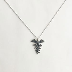 May include: A silver-toned necklace featuring a pendant shaped like a stylized spine with bat wings at the top. The pendant has a detailed, textured design. The chain is delicate and silver, set against a white background.