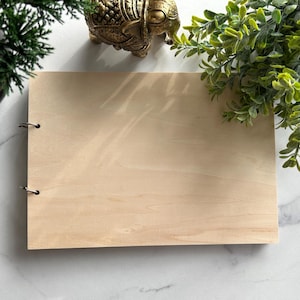 Puede incluir: Un cuaderno de madera de color claro con anillas metálicas. El cuaderno es rectangular y tiene una superficie lisa sin terminar. Plantas decorativas y una estatua de elefante dorado están en el fondo.