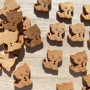 Puede incluir: Una colección de botones de madera de color marrón claro con forma de gato de dibujos animados. Cada botón tiene dos agujeros para coser y una cara sonriente. Los botones están dispersos sobre una superficie clara.