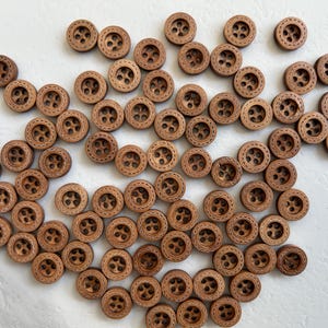 May include: A large group of brown wooden buttons with a stitched detail around the edge. The buttons are arranged in a random pattern on a white surface.