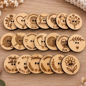 Pode incluir: Uma coleção de botões de madeira clara de formato redondo com desenhos gravados a laser, incluindo flores, corações, ovelhas, novelos de lã, máquinas de costura, pegadas e textos como Handmade with Love, cada um com dois orifícios.