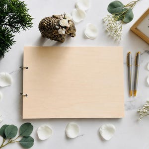 Puede incluir: Un libro de visitas de madera con encuadernación plateada, dos bolígrafos y un pequeño letrero enmarcado que dice "Please Share Your Wishes". La escena está decorada con pétalos de flores blancas, vegetación y una estatua decorativa de elefante.
