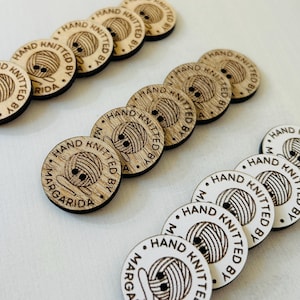 Könnte beinhalten: Eine Sammlung runder, heller Holzknöpfe mit einem lasergravierten Motiv eines Wollknäuels. Jeder Knopf ist am Rand mit dem Text "HAND KNITTED BY MARGARIDA" versehen und verfügt über zwei kleine Löcher zum Annähen an Kleidungsstücke.