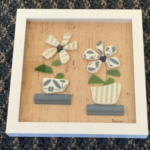 May include: A framed artwork featuring two stylized flower arrangements. The flowers are composed of light blue and white ceramic pieces, with green leaves and dark blue centers. The pots are in light blue and white, set on a gray base, against a cork background.