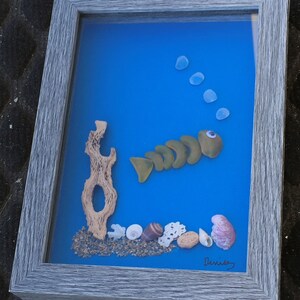 May include: Framed artwork featuring a whimsical fish made of green sea glass, with a driftwood accent and seashells on a blue background. The gray frame surrounds the ocean-themed art piece.