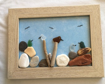 May include: Framed artwork featuring a beach scene with seagulls flying overhead. The artwork is made with sea glass and stones, depicting birds perched on driftwood and rocks. The piece is signed "Denise".