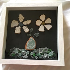 May include: A framed artwork featuring a flower arrangement made of white and green sea glass. The flowers are in a vase made of sea glass and are set against a black background. The artwork is signed "Denise".