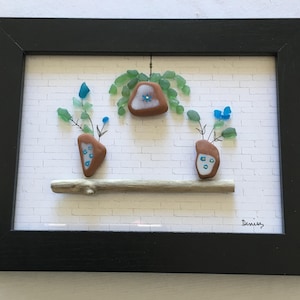 May include: A framed artwork featuring three small vases made of sea glass and driftwood. The vases are decorated with blue flowers and are arranged on a white brick wall background. The artwork is signed "Denise".
