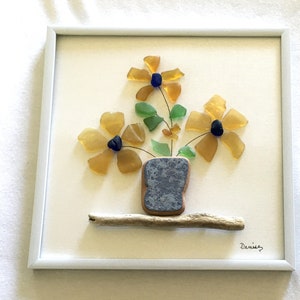 May include: A framed artwork featuring a floral arrangement made of sea glass. The flowers are yellow with blue centers, green leaves, and are arranged in a blue vase. The artwork is mounted on a cream-colored background and includes a driftwood shelf.