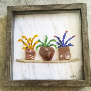 May include: Framed artwork featuring three potted plants made of colorful glass pieces. The plants are arranged on a wooden shelf against a marble-patterned background. The frame is a weathered gray color.